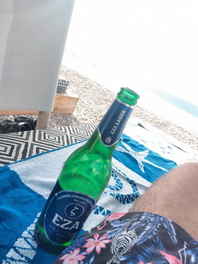 A green glass bottle of EZA Lager, partially obscured by a person's leg, sits on a beach towel. The towel is blue and white with a pattern of stars. The beach is covered in pebbles. In the background, the sea is visible under a bright sky. The scene appears to be a relaxed setting, likely during a vacation, given the beach environment and casual attire.
