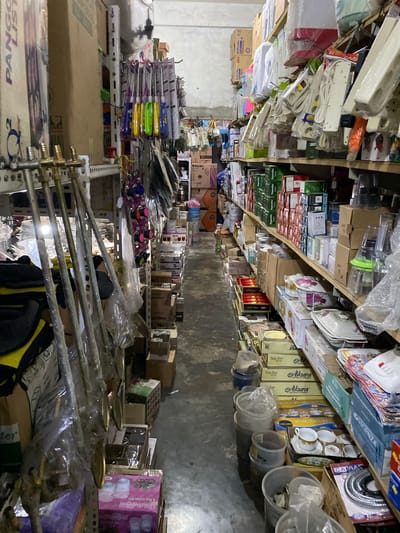 This image shows a cluttered storeroom filled with various household items. The shelves are packed with boxes of kitchenware, appliances, and other packaged goods. Among the visible items are multiple sets of power strips, tea sets, and what appear to be decorative plates or saucers. Some items are still in their original packaging, while others are loosely stored.
The scene is dimly lit, suggesting an indoor environment that is not actively being used or is perhaps a storage area for a shop or warehouse. The items are densely arranged, creating a sense of disarray. The visible text includes brand names like "Miyako" and "Hyperlite," as well as product descriptions like "Tea Set" and "6 Pcs Glass Set." The overall impression is of a storage space overflowing with merchandise, possibly awaiting sale or distribution. The location context of Ajuen, Indonesia, suggests this could be a typical storeroom for a local business in that region. There are no people visible in the image, and the focus is entirely on the objects and the setting.