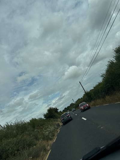 The image shows a view from a vehicle driving on a rural road in New Ross Municipal District, Ireland. The sky is filled with heavy, gray clouds, with patches of blue sky visible. Three cars are on the road ahead, a dark gray hatchback in the left lane, followed by a red hatchback in the right lane. A fourth car, a darker color, is visible further ahead. The road is asphalt, with a yellow line marking the edge on the left and a dashed white line separating the lanes. Lush green vegetation, including bushes and trees, lines the right side of the road, with taller trees in the distance. Utility poles with overhead wires run along the right side of the road. The scene appears to be during daylight hours, likely overcast.