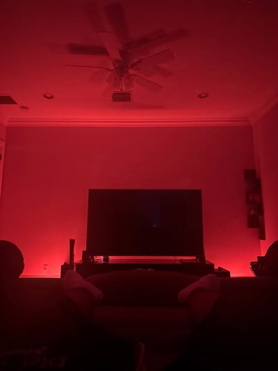 A living room in Parkland, United States of America, is depicted, illuminated by a pervasive red light source. The perspective is from a seated position, looking towards a prominent television setup. In the foreground, the lower legs and feet of an individual, wearing white socks, are visible, resting on a dark surface, likely a sofa or lounge chair. Dark, indistinct shapes on the extreme left and right edges suggest additional seating.
Centrally located against the far wall is a large, dark-screened television, positioned on a dark media console. To the left of the television, a vertical white object, resembling a gaming console, rests on the media console. A white electrical outlet is visible on the lower left section of the wall. Along the base of the wall beneath the television and console, a strip of red light is visible, contributing to the room's illumination. To the right of the television, a vertical arrangement of dark, rectangular shelves or wall decor is mounted on the wall.
The ceiling features a multi-bladed ceiling fan, two recessed light fixtures, and a rectangular vent. White crown molding lines the intersection of the walls and ceiling. The red lighting casts subtle shadows from the ceiling fan.