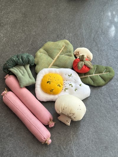 A flat lay image shows a collection of fabric toy food items arranged on a textured, dark gray surface, possibly a stone countertop or table. The toys include two pink sausage-like objects tied at the ends with twine, a green broccoli floret, a fried egg with a yellow yolk and white "white," a small red tomato with a green stem and leaves, a cream-colored mushroom with a tan stem, and two green fabric leaves. One leaf has prominent white stitching to resemble veins, and another, larger leaf shape is also visible. The overall aesthetic is rustic and handmade. The image is a close-up shot, with the top half of the frame occupied by the gray surface. There are no people or animals present in the image. The location in Chornomorske, Ukraine is not discernible from the image itself.