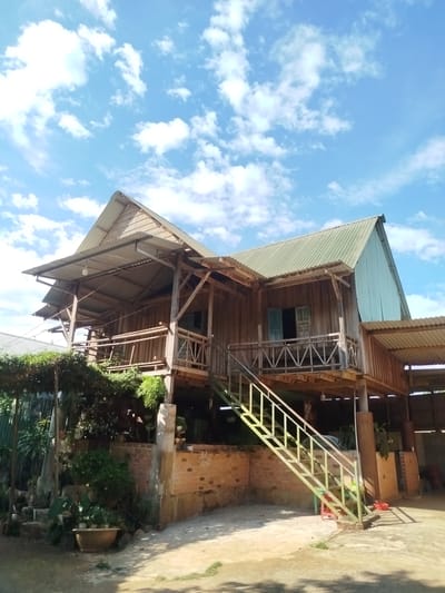 The image shows a wooden house elevated on stilts, typical of structures found in An Phu Ward, Vietnam. The house is constructed primarily of wood with corrugated metal roofing. A set of metal stairs leads up to a balcony on the front of the house, with a second balcony visible on the left side. The area beneath the house appears to be open, possibly used for storage or parking. Vegetation grows along the base of the structure and to the left. The sky is visible in the background, featuring a combination of blue sky and scattered clouds. The foreground shows a paved area with some vegetation. No people are visible in the image.