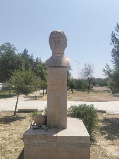 A bronze bust of a young man with dark, neatly combed hair, is mounted on a stone pedestal. The bust is positioned centrally in the frame, facing forward. The pedestal has inscriptions in a Cyrillic script, reading "ТАЛБАК АДАЕВ" (Talbak Adaev) vertically, with a stylized ear of wheat beneath the name. Below the inscribed section of the pedestal, there is a large, broken clay pot with several fragments scattered around its base.
The monument stands on a concrete base in an outdoor, arid environment. The ground is covered with dry grass and sparse vegetation. To the left and right of the monument, young coniferous trees are planted. In the background, there are more trees, a street light, and a low-rise building with a brown, sloped roof. The sky is clear and blue. The overall scene suggests a monument in a public park or memorial area within the city of Danghara, Tajikistan. The condition of the broken pot indicates possible vandalism or natural decay.