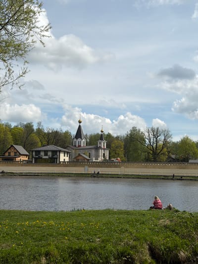 The image shows a view in the city of Pavlovo, Russia. A pond or lake stretches across the middle ground of the image, bordered by a stone wall and buildings on one side, and a grassy bank with yellow flowers on the other. A person in a pink coat is near the edge of the grassy bank. Behind the stone wall and buildings are trees, and two church steeples are visible, with a sky full of clouds above.