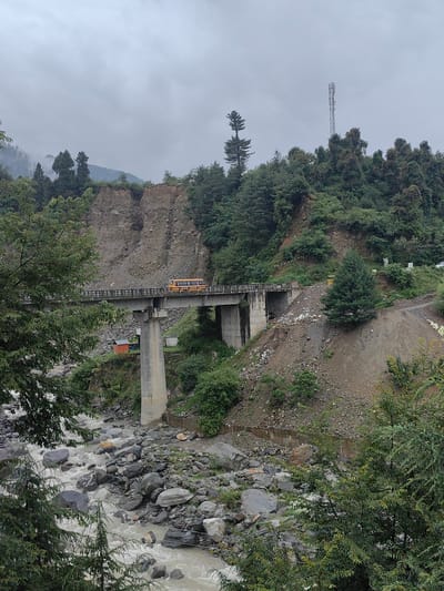 An overcast day frames a scenic view in Aleo, India, dominated by a large concrete bridge spanning a rocky river. A yellow school bus is visible on the bridge, traveling from left to right. The surrounding landscape is mountainous and heavily forested, with steep, eroded slopes and dense evergreen trees. A communication tower stands tall on a distant ridge. The river below is wide and turbulent, with numerous large rocks scattered throughout its course. There are no visible people in the image, but there is evidence of a small orange structure near the bridge abutment. The overall impression is one of a remote, natural environment with signs of infrastructure.