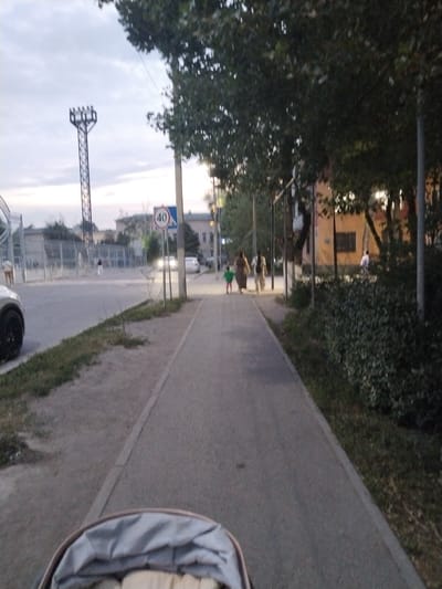 The image depicts a street scene in Ötegen Batyr, Kazakhstan, likely during dusk or early evening, as indicated by the subdued lighting and the illuminated streetlights. The foreground shows the top of a baby stroller, suggesting the perspective of someone pushing it. A paved sidewalk runs alongside a road, which has a speed limit sign indicating 40.

Several individuals are visible. In the middle ground, a woman wearing a dark top and lighter pants is walking with a young child in a green outfit. Behind them, another person in a lighter outfit is also walking. Further down the sidewalk, a person in a white top and dark pants is walking away from the camera. On the road, two cars are approaching, their headlights on, illuminating the scene.

The background features trees with dense foliage lining the sidewalk. Beyond the trees, there are buildings, some appearing to be residential, and a tall, industrial-looking structure with multiple lights at its top. A chain-link fence is also visible, along with a power line stretching across the sky. The weather appears to be overcast. The overall mood of the image is tranquil, capturing an ordinary moment of daily life in a residential area.