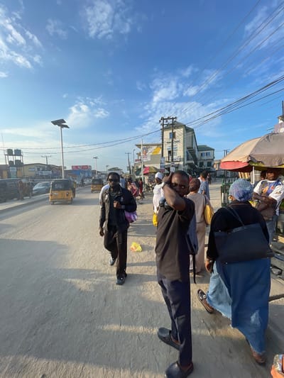 The image depicts a bustling street scene in Lagos, Nigeria, likely during the late afternoon given the long shadows cast across the pavement. The sky is a clear blue with scattered white clouds. The scene is oriented vertically, with the ground appearing at the top of the image.
Several people are visible, some walking and others gathered near market stalls. In the foreground, a person wearing a black t-shirt and dark pants is looking towards the camera, with headphones around their neck. Further back, a group of people are seated under a canopy, possibly at a market stall or a waiting area. One individual wears a blue cap with a patterned design. Another person in the mid-ground is dressed in a dark jacket and light-colored trousers, with a backpack slung over their shoulder. In the background, several vehicles, including a yellow auto-rickshaw, are on the road, suggesting moderate traffic. Buildings line the street, with signage indicating businesses and shops.
The overall atmosphere appears lively and dynamic, typical of a busy urban environment. The sunlight indicates it is daytime. The presence of auto-rickshaws and the architectural style of the buildings are strong cues for the location being Lagos, Nigeria. Visible text includes signs on buildings and possibly advertisements on vehicles, though the text is largely illegible due to resolution and orientation.