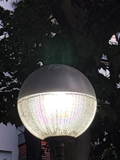 The image shows a close-up view of an outdoor lamp with a translucent, textured globe. The lamp is illuminated from within, casting a bright white light that diffuses through the globe, creating a rainbow effect due to the texture. The top of the lamp is a solid, dark gray cap. The lamp is mounted on a pole, which is partially visible at the bottom.

The background is dark and out of focus, suggesting it is nighttime or a dimly lit area. Silhouetted against the sky are the dark branches and leaves of trees. In the lower left portion of the image, a small section of a white building with some horizontal striped elements is visible, possibly indicating it is a building adjacent to the lamp.

The image does not contain any people or visible actions. The scene appears to be an urban streetlamp, likely in a park or along a pathway in the city of Coslada, Spain. The overall impression is one of functional outdoor lighting against a natural and architectural backdrop.
