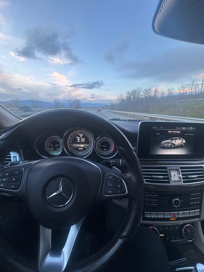 The image shows the interior of a Mercedes-Benz car, focusing on the dashboard and steering wheel. The car is driving on a road, with a view of the sky and trees outside. The dashboard displays information such as speed (113 km/h), engine temperature (95°C), and outside temperature (12.0°C). The central display shows "Seat Settings" and indicates that the time is 18:34. The location is given as VIVACO in NW Bulgaria.