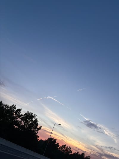 The image depicts a dusky sky over a road in Mirošovice, Czechia. The sky transitions from a deep blue at the top to hues of pink and orange near the horizon, indicative of sunset. Wispy clouds are scattered across the sky, with two prominent contrails crossing each other in the upper right quadrant. Silhouetted trees line the left side of the frame, partially obscuring the view of the road below, which features a concrete barrier. A single street lamp is visible on the right side of the road, its light directed downwards. The scene is devoid of any people or vehicles.
