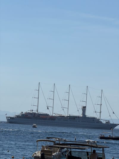 The image shows a tall ship with multiple masts anchored in the sea. The ship is large and appears to be a cruise ship with a tall, slender hull. The masts are equipped with rigging, but the sails are not visible. The sea is calm, and there is a hint of land in the distance. Several smaller boats are nearby. The image was taken from a slightly elevated position, likely from another boat or a nearby structure. Pogerola is a village high above Amalfi. The boat is too far to be photographed from that village.