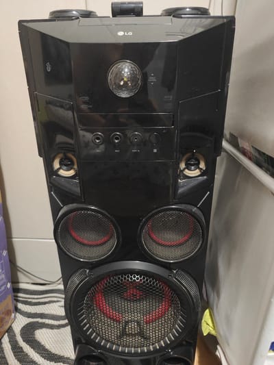 An LG audio system speaker tower is pictured in a domestic setting, possibly a living room. The tower is tall and black, with multiple speaker grills. The top of the tower features a rotating disco ball light and various control buttons and ports, including USB inputs and microphone jacks. Two medium-sized speakers are positioned above a larger, main speaker at the bottom. The speaker is placed on a rug with a black and white striped pattern, and the background shows a neutral wall and a portion of a white curtain. The image does not contain any elements that indicate a location within the city of Parabel, Russia.