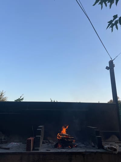 The image depicts an outdoor scene with a clear blue sky. In the foreground, a barbecue pit is visible, with a fire burning inside. The pit appears to be constructed of bricks and metal. There are some unburnt logs and bricks stacked nearby. On the left side of the image, there are tree branches with leaves hanging down. In the upper portion of the frame, a wooden beam with a light fixture attached extends horizontally across the sky, and some wires are strung. The lighting suggests it is daytime, possibly late afternoon, given the bright sky. The scene appears to be in a residential or recreational outdoor area.