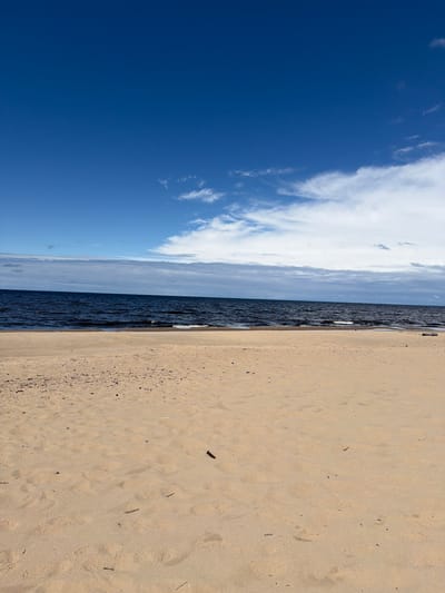A bright, sunny day at the beach in Lilaste, Latvia, is captured in this vertical, eye-level shot. The scene is divided into three distinct horizontal bands. The top portion of the image is dominated by a vast expanse of clear, deep blue sky, with a few wispy white clouds scattered across it. Below the sky, a dark blue body of water, likely the Baltic Sea, stretches across the frame. The water exhibits gentle ripples, indicative of a light breeze, and the horizon is marked by a thin line of pale clouds. The foreground of the image is a sandy beach, light beige in color, with subtle undulations and scattered small pieces of driftwood and debris. The sand appears dry and undisturbed, suggesting it is not a heavily trafficked area at this moment. There are no people or animals visible in the frame, and the overall impression is one of peaceful solitude and natural beauty.