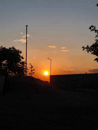This is a vertical, outdoor, full-shot photograph taken at sunset in Benkovski, Bulgaria. The sky is a gradient of soft blues and oranges, with scattered clouds. In the foreground, a dark silhouette of a grassy area with trees and structures creates a stark contrast against the vibrant sky. A tall, thin pole with a cross-like fixture stands prominently on the left side of the image. A large, dark building or wall occupies the right side, with a streetlamp visible near its center. The sun, a bright orb, is setting behind a gentle hill in the middle-ground, casting a warm glow on the clouds. Silhouetted trees with leafy branches frame the top right and left corners of the image. The overall mood is serene and atmospheric, capturing the tranquil beauty of twilight in an urban setting.