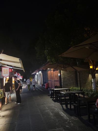 This is a night scene of a pedestrian street in Wuhou District, China. The street is paved with stone slabs and lined with shops on both sides. On the left, there are several shops with awnings and signs, one of which has Chinese characters. People are walking along the street, some are looking at the shops, and others are walking away from the camera. On the right, there are outdoor tables and chairs under umbrellas, suggesting a dining or resting area. The buildings on the right are made of brick and have traditional Chinese architectural features. Trees line the right side of the street, and some have yellow lights. The overall atmosphere is lively and commercial, typical of a night market or shopping street.