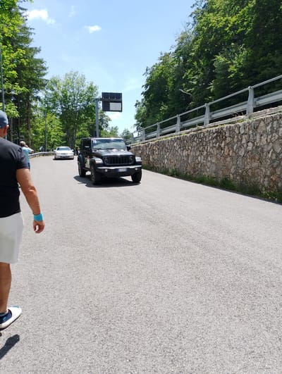 The image depicts a winding asphalt road bordered by a stone wall and lush green trees on a sunny day. In the foreground, a person wearing a black t-shirt and white shorts is walking away from the camera, with a blue wristband visible on their left arm. Further down the road, a black Jeep Wrangler with the license plate "CY-ST 05E" is driving away. To the left of the Jeep, a white sedan is also seen on the road. A traffic sign is visible on the left side of the road, and a metal guardrail runs along the top of the stone wall on the right. The environment suggests a scenic route, possibly in a mountainous or hilly area. The city of Rieti, Italy, is known for its surrounding natural beauty and winding roads that lead to mountainous areas, making this scene consistent with a location in or around Rieti.
