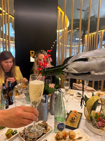 The image shows an indoor scene in what appears to be a restaurant. Champagne is being poured from a green bottle held within a grey cooler into a champagne flute. The table is set with various dishes including sushi, raw fish, and shellfish. There are multiple glasses and bottles, including a bottle of Font d'Or mineral water. Two women are visible, one seated and the other partially visible behind a floral decoration. The hand holding the champagne flute is also visible. The environment suggests a modern restaurant interior with a decorative metal screen in the background. There is no information indicating whether it is Torreforta, Spain.