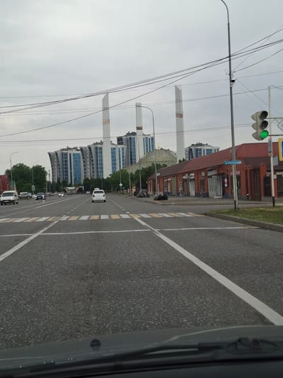 The image shows a street scene in Argun, Russia, with a pedestrian crossing in the foreground. In the background, there are modern apartment buildings with distinctive curved facades and white tower-like structures. A red brick building with a sign in Cyrillic script that reads "Н. КРУПСКОЙ" is visible on the right side of the street. A traffic light with a green signal is also present. Several cars are driving on the road, including a white van, a black sedan, and a white hatchback. The sky is overcast, and the overall atmosphere is that of a cloudy day.