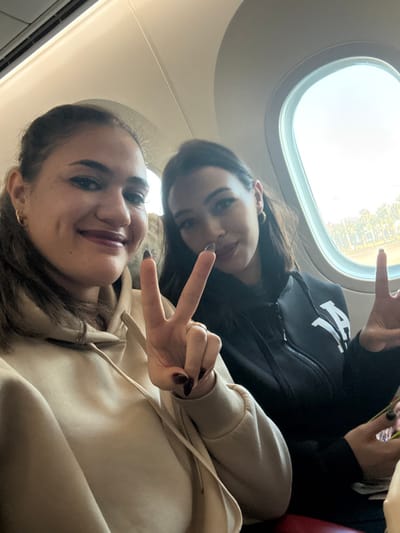 The image displays two adult females seated within an aircraft cabin, located in Nouaceur, Morocco. Through an oval window on the right, an airport environment is visible, including a tarmac and green vegetation under bright daylight. The woman on the left, with dark hair tied back, wears a light beige hooded sweatshirt. She is smiling, revealing dimples and dark lipstick. Her right hand is raised in a peace sign, with dark-painted fingernails and a gold-colored ring on her ring finger. A gold hoop earring is visible in her left ear. The woman on the right, with dark hair, wears a black zip-up jacket featuring a white "LA" style logo on the chest. She is also smiling and holds her left hand up in a peace sign, displaying dark-painted fingernails. A gold hoop earring is visible in her right ear. Both individuals are looking towards the camera. A red seat portion is visible in the lower right corner.