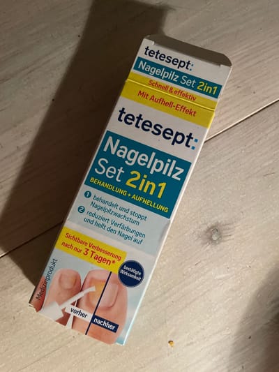 This image shows a product box for "tetesept Nagelpilz Set 2in1" (tetesept Nail Fungus Set 2in1). The box is positioned on a light-colored, wooden surface, possibly a floor or table, with visible wood grain. A shadow is cast to the left of the box, suggesting the light source is to the right. The product is for treating nail fungus and claims to be fast and effective with a brightening effect. It also highlights visible improvement after just 3 days and confirmed effectiveness. The packaging features graphics of toes, illustrating a "before" and "after" application of the product. The product itself appears to be a medical product.
The image does not contain any information or visual cues that would indicate its location in the city of Oldenburg in Holstein, Germany.