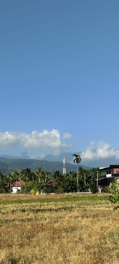 The image depicts a rural landscape in Bateiiliek, Indonesia. In the foreground, a field of dry, golden grass stretches across the frame. Behind the field, a line of lush green trees and palm fronds obscures a cluster of buildings. To the left, a white house with a red roof is visible. Further to the right, a taller building with dark walls is partially visible. In the mid-ground, a red and white radio tower stands tall against a backdrop of rolling green hills that fade into the distance, disappearing into a hazy, blue sky dotted with scattered white clouds. The overall scene is calm and serene, with a sense of open space and natural beauty.