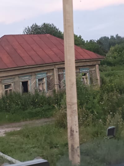 The image depicts an old, weathered wooden house with a red metal roof, situated in a rural setting near the village of Махалино, Russia. The house appears to be abandoned or in disrepair, with some windows boarded up or broken. The surrounding environment is overgrown with tall grass and vegetation, suggesting a lack of recent human activity. A concrete utility pole stands prominently in the foreground, obscuring part of the house and its surroundings. The sky is overcast with a light, diffused glow, indicating either early morning or late evening. The overall impression is one of neglect and decay in a rural landscape.