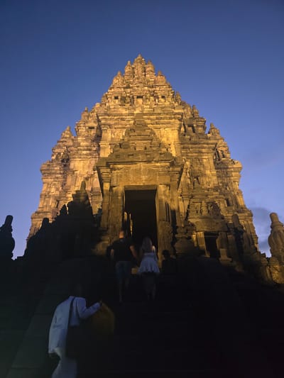 Captured at night in Bokoharjo, Indonesia, the image depicts a multi-tiered stone structure, illuminated in a warm golden glow against a twilight sky. The structure, likely a temple, is built of stacked stone blocks, with intricate details and carvings visible on its surface. The lower portion of the image is obscured by shadow, with several figures visible on the right side, including individuals with light-colored clothing.