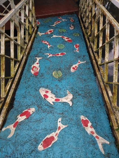 The image displays a pedestrian pathway or bridge deck, located in Elverum, Norway, viewed from an elevated perspective. The central surface of the pathway is painted a mottled, textured blue, intended to simulate water. Emblazoned on this blue surface are approximately fifteen to twenty stylized koi fish, depicted in white with irregular red-orange blotches, oriented in various directions to suggest movement. Several green lily pads are also painted intermittently among the fish.
The pathway is bordered on both sides by identical metal railings. These railings are off-white or light gray, heavily weathered, exhibiting extensive rust, paint degradation, and patches of green biological growth consistent with moss or algae. They are constructed with a grid-like pattern of intersecting vertical and horizontal bars. At the upper termination of the blue pathway, a reddish-brown tiled or paved surface is visible. Beyond the railings, a dark, reflective surface, consistent with water, is present, reflecting overhead light. No individuals or active interactions are depicted within the scene.