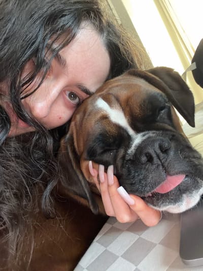 A close-up photograph shows a woman lying down with a brown and white Boxer dog. The woman's face is partially visible on the left side of the frame, with dark hair obscuring part of her face. Her right eye is visible. The Boxer dog is positioned on top of her, with its face close to the camera. The dog's tongue is sticking out slightly. The woman's hand, with long, manicured nails, is holding the dog's head. The background appears to be a light-colored surface with a gray and white geometric pattern, possibly a pillow or blanket. The photograph was not taken in the city of Lissone, Italy.