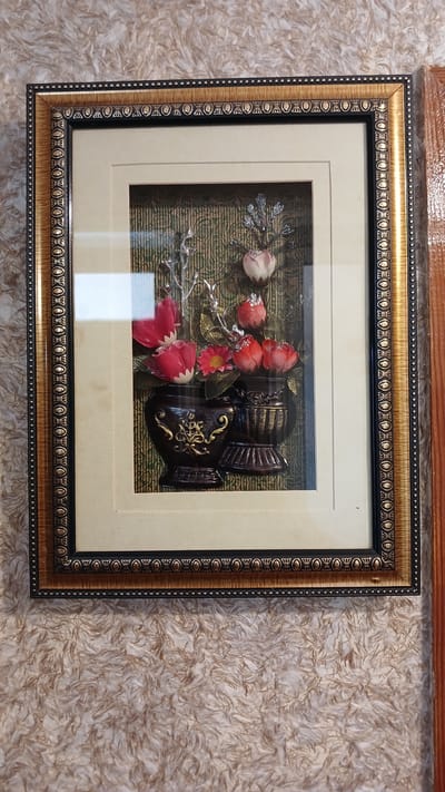 A framed 3D artwork of flowers in vases hangs on a textured, light-colored wall. The artwork features red and pink flowers with metallic stems and leaves, displayed in two dark brown vases with gold accents. The frame is ornate with multiple decorative borders in gold and black. The wall has a fluffy, shaggy texture. The image shows a section of a wooden door frame on the right side. The location is not specified in the image.