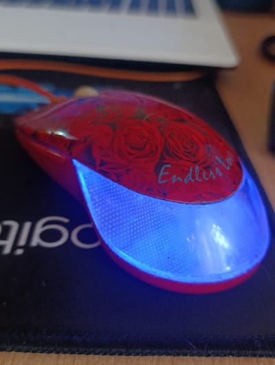 The image displays a close-up of a red computer mouse with a floral design of red roses. The word "Endless love" is inscribed in a light blue, cursive font on the top surface of the mouse, over the rose pattern. The mouse has a blue LED light that illuminates the front portion and the scroll wheel area, creating a glowing effect. The mouse is positioned on a black mousepad with a visible logo of "Logitech" in white text. In the background, a portion of a white laptop or keyboard is visible, along with a wooden surface below the mousepad. The lighting suggests an indoor environment, likely an office or home desk setup. There is no indication of the time of day or weather. The overall mood conveyed is personal and perhaps sentimental due to the "Endless love" inscription and the rose design. The context provided is Yenakiieve, Ukraine, but there are no specific visual cues within the image to confirm this location.