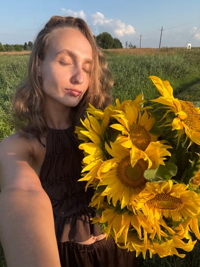 A young woman with light brown hair is holding a bouquet of sunflowers. She is looking at the camera with her eyes closed and her lips slightly puckered. She is wearing a dark brown halter top with ruffles. The background is a field of green grass with trees and power lines in the distance. The sky is blue with some white clouds. The scene appears to be in Aksaitovo, Russia.
