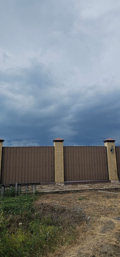 The media file displays a brown metal fence with decorative brick pillars topped with dark caps. A light fixture is visible on one of the pillars. The fence is bordered by a stone-clad base. To the right of the fence, there is dry, tall grass and some green vegetation. The sky above is a mix of blue and white clouds, suggesting daytime with potentially changing weather. The scene appears to be outdoors, possibly in a rural or suburban setting, given the vegetation and fencing. There are no people visible in the image. The overall impression is one of an exterior view of a property boundary. The location context indicates the scene is in Овощной, Russia.