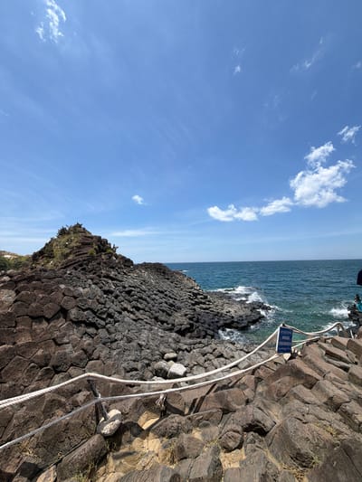 The image depicts a scenic view of the basalt columns of Gành Đá Đĩa in the commune of An Ninh Đông, district of Tuy An, in the city of Phú Lương, Vietnam. The foreground is dominated by a rugged expanse of dark, hexagonal basalt rock formations, forming a natural staircase-like pattern. A thick white rope, acting as a safety barrier, is strung across several wooden posts, guiding visitors through the rocky terrain. Beyond the rocks, a turquoise ocean stretches to the horizon, with gentle waves lapping against the shore, creating white foam. The sky is a vibrant blue, with scattered white clouds adding depth and texture to the scene. A small blue sign with white lettering is visible near the rope barrier, likely providing information or safety instructions. The overall atmosphere is one of natural beauty and geological wonder.