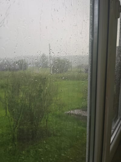 The image captures a view through a window with raindrops on the glass. The exterior scene is blurred by the rain and water droplets, but it appears to be a green, outdoor landscape, possibly a field or park, with some trees visible. The sky is overcast and grey, indicating rainy weather. On the right side of the image, a portion of the window frame is visible, made of white material. There are no people or specific landmarks that would identify the location within Малая Уса, Russia. The overall impression is one of a calm, rainy day viewed from indoors.
