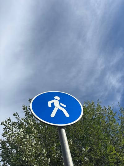 A circular blue sign with a white pictogram of a person running is mounted on a metal pole. The sign is positioned to the right of the frame, partially obscured by lush green foliage from a tree. The background is a pale blue sky with wispy, white clouds, suggesting a partly cloudy day. The lighting indicates it's daytime, likely midday or early afternoon, with the sun illuminating the scene from the side. The image is taken from a low angle, looking upwards towards the sky. The overall impression is one of an outdoor setting, possibly a park, pathway, or recreational area in Vsevolodovo, Russia, where the sign indicates a designated running path or zone. There is no visible text on the sign or in the surrounding environment.