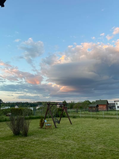 This landscape image depicts a serene rural or suburban backyard scene in Zaķumuiža, Latvia, likely captured during sunset or sunrise. The sky occupies a significant portion of the frame, displaying a dramatic interplay of light blue areas and diverse cloud formations. Dark grey, heavy clouds are interspersed with lighter clouds illuminated by warm, peachy, and orange tones, characteristic of golden hour.
The foreground and midground consist of a wide expanse of green lawn. Centered in the midground is a wooden A-frame swing set with a suspended hammock-style seat. Adjacent to the swing set are two light blue and white striped deck chairs. A large, dark bush with bare branches is visible closer to the foreground.
In the background, several structures are visible, including a house with a white exterior and a dark roof, along with multiple smaller wooden outbuildings or sheds featuring green or brown roofs. Fences can be seen dividing properties. A dense line of trees forms the horizon line. No people are present in the image. The scene conveys a sense of peacefulness and natural spaciousness under a dynamic sky. There is no visible text.