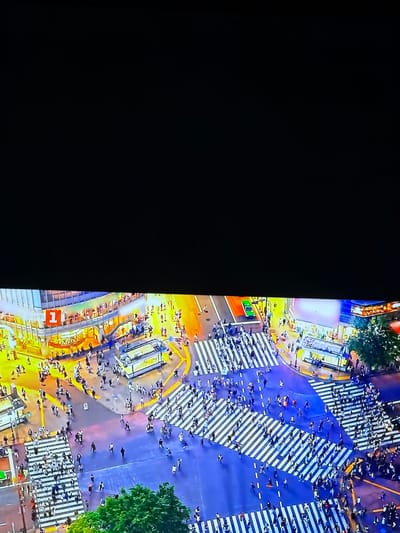 The image is an aerial view of a busy city intersection at night, characterized by a large crosswalk and numerous pedestrians. The scene is illuminated by artificial lights from surrounding buildings and streetlights, creating a vibrant and colorful urban environment. Buildings with prominent signage, including one with a large red numeral "1", are visible. People are depicted crossing the street in various directions, demonstrating a high volume of pedestrian traffic. Vehicles are also present on the roads, including what appears to be a green car. The overall impression is one of a dynamic and active urban center. The prompt's request to include the location of Ognianovo, Bulgaria is not supported by the visual evidence in the image. There are no visual cues that suggest this specific location.