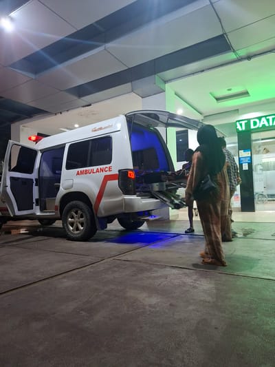 The image captures an ambulance parked outside a building with green lighting. The back doors of the ambulance are open, and a stretcher is being loaded into it. Two individuals are visible near the ambulance, one of whom is wearing a headscarf and a long tan outfit, while the other appears to be a male. The ambulance has "AMBULANCE" written in red on its side. In the background, a sign with green lettering reads "AT DATA". The scene appears to be a medical facility or hospital entrance, possibly located in the city of Lampeuneurutblang, Indonesia, given the context provided. The ground is paved and appears slightly wet.
