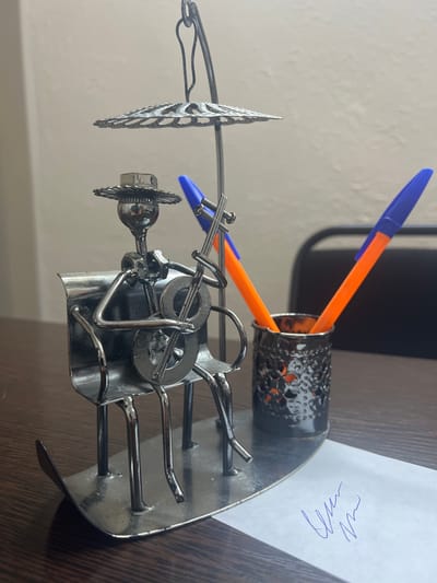 This media file features a desk ornament crafted from metal, depicting a stylized figure playing a musical instrument. The figure is seated and appears to be holding a guitar-like instrument. The ornament is placed on a wooden desk, with a portion of a white paper featuring blue scribbled writing visible on the right side. Two pens, one blue and one orange, are resting in a metallic cup, also part of the ornament. The background is out of focus, showing a light-colored wall. The scene appears to be indoors, in a brightly lit environment. The overall impression is that of a decorative desk accessory. The location context provided is Satbayev, Kazakhstan.