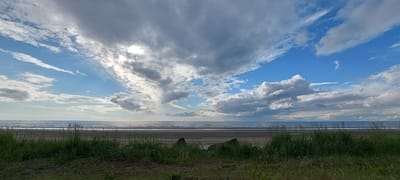 The image depicts a natural coastal scene. In the foreground, tall green grass grows along the edge of a sandy beach. Beyond the beach, the calm sea stretches to the horizon, meeting a sky filled with a dramatic display of clouds. The clouds vary in density and color, with some appearing bright white and others a darker gray, suggesting dynamic weather conditions. Sunlight breaks through in some areas, creating illuminated patches in the sky. The overall impression is of an expansive, somewhat overcast but bright, natural landscape. The location is identified as Aleksandrovsk-Sakhalinskiy, Russia.