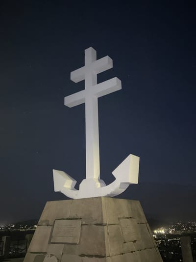A white anchor and cross sculpture is mounted on a concrete base. The sky is dark, and lights from the town are visible in the background. This sculpture is the Gourock memorial to Free French forces.