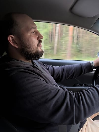A medium shot shows a man with a beard and a shaved head driving a car, seen from the side. He is looking straight ahead through the windshield, his hands on the steering wheel. He is wearing a dark hooded sweatshirt and a seatbelt. The background outside the car window shows a blurred view of trees, suggesting a rural or forested area. The car interior is dark. The man's facial expression is neutral.