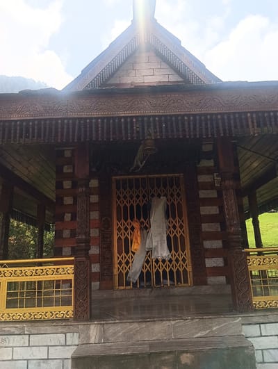 The image shows the exterior of a wooden temple structure in Khakhnal, India. The temple features intricate wood carvings on the roof and pillars. A golden, collapsible gate guards the entrance, adorned with two pieces of cloth, one orange and one white. A small bell hangs above the entrance. The temple is set against a backdrop of green hills and a bright, cloudy sky, with the sun's rays creating a lens flare effect at the top center of the image. The ground in front of the temple is paved with stone.