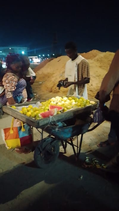 At night in Effurun, Nigeria, a street vendor stands behind a cart displaying a collection of yellow fruit, most likely oranges and lemons, with a slice of watermelon. The cart is an old blue wheelbarrow. The vendor is a young man in a light-colored shirt, handling something in his hands. A woman with a patterned dress and head covering is browsing the fruit with another person. A sand pile is in the background. There is a yellow, red, and blue tote bag hanging on the cart.