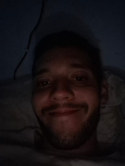The image depicts a young man lying in bed, captured from the chest up. He is wearing a dark shirt. He is smiling and looking directly at the camera. The image appears to be taken in low-light conditions, as the environment is mostly dark. The man has short dark hair, a goatee, and is wearing a small circular earring in his right ear. The location of this image is in Angra dos Reis, Brazil, based on the provided coordinates.