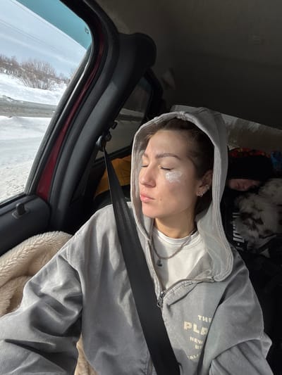 An image captured inside a vehicle depicts two individuals and a winter landscape. In the foreground, a young adult female is seated, secured by a black seatbelt across her chest. She wears a light grey hooded zip-up jacket over a white top and a silver chain necklace with a dark pendant. Her eyes are closed, and a visible dollop of white cream is present on her left cheekbone. Multiple small hoop earrings adorn her left ear. To her left, a cream-colored, fuzzy textile is visible. In the background, to the right of the primary subject, a second individual, possibly a child, is partially visible, appearing to be asleep. This individual wears a black beanie, and a white and brown patterned object, possibly a blanket or animal fur, rests near them. Behind this second individual, a silver, reflective material is present, likely a sunshade or insulation. Through the car window on the left, a snow-covered outdoor environment is visible, featuring bare trees or shrubs on a gentle slope, under a bright, clear sky. A metal guardrail is partially discernible along the roadside. The scene is located within the city of Ustinovo, Russia.