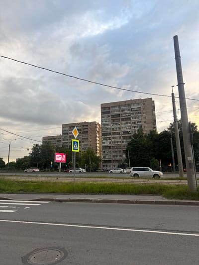 The image depicts an urban street scene in Kokkorevo, Russia, with a focus on a multi-lane road and residential buildings. The sky is overcast with clouds. On the left side of the road, a pedestrian crossing is marked by white lines. A large digital billboard displaying pink advertisements is visible in the middle ground. Adjacent to the billboard, traffic signs, including a pedestrian crossing sign and a diamond-shaped warning sign, are mounted on a pole. Several cars are driving on the road, including a silver SUV on the right. Behind the road, a line of trees separates the road from a row of tall, multi-story apartment buildings with numerous windows. Two large concrete utility poles stand on the right side of the image, one prominently in the foreground. Power lines are strung across the sky. The overall impression is of a typical day in a Russian city, with infrastructure and residential life coexisting.