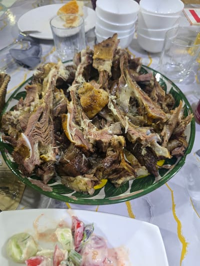 The image depicts a close-up, eye-level view of a table setting with a large platter of meat and a side dish of salad, likely from a meal in Guzar, Uzbekistan. The central focus is a green-rimmed ceramic platter overflowing with pieces of cooked meat, appearing to be lamb or mutton. The meat is cut into various sizes, with bones visible, and has a glistening, slightly charred appearance, suggesting it has been roasted or grilled. Some white shredded cheese or fat is sprinkled over the meat. In the foreground, a white rectangular plate holds a portion of a salad, composed of chopped tomatoes, cucumbers, and possibly onions, mixed with a creamy pinkish dressing. The table is covered with a white tablecloth featuring abstract yellow and green patterns. To the left of the meat platter, a white plate with a piece of bread is visible, along with a glass of water and a folded gray napkin. In the background, stacks of white ceramic bowls are arranged, and another glass of water is present. The overall impression is of a home-cooked meal, possibly a celebration or gathering, set within a domestic or casual dining environment in Guzar.